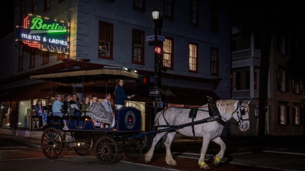 A white horse pulls a blue carriage filled with people during a nighttime city tour under a neon sign that reads “Berlin’s Men’s & Ladies.
